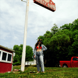 Album cover of a woman standing in grass in jeans under a sign and near a red truck