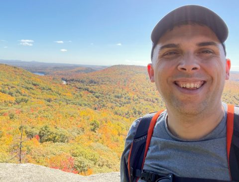 Close up of Jason Clarke's face standing on a mountain with orange and yellow foliage behind him