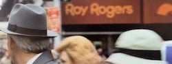 A man waits for a cab in a crowded ew York City street with a Roy Rogers restaurant sign visible in the background