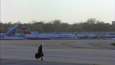 A man walks across an airport runway with a TWA jet visible in the background.